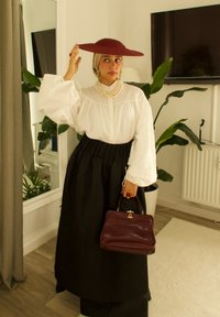 Czerwona kapelusz z szerokim rondem, biała bluzka z bufkami, czarna spódnica i naszyjnik z pereł. Trzyma bordową torebkę. Wnętrze z roślinami.