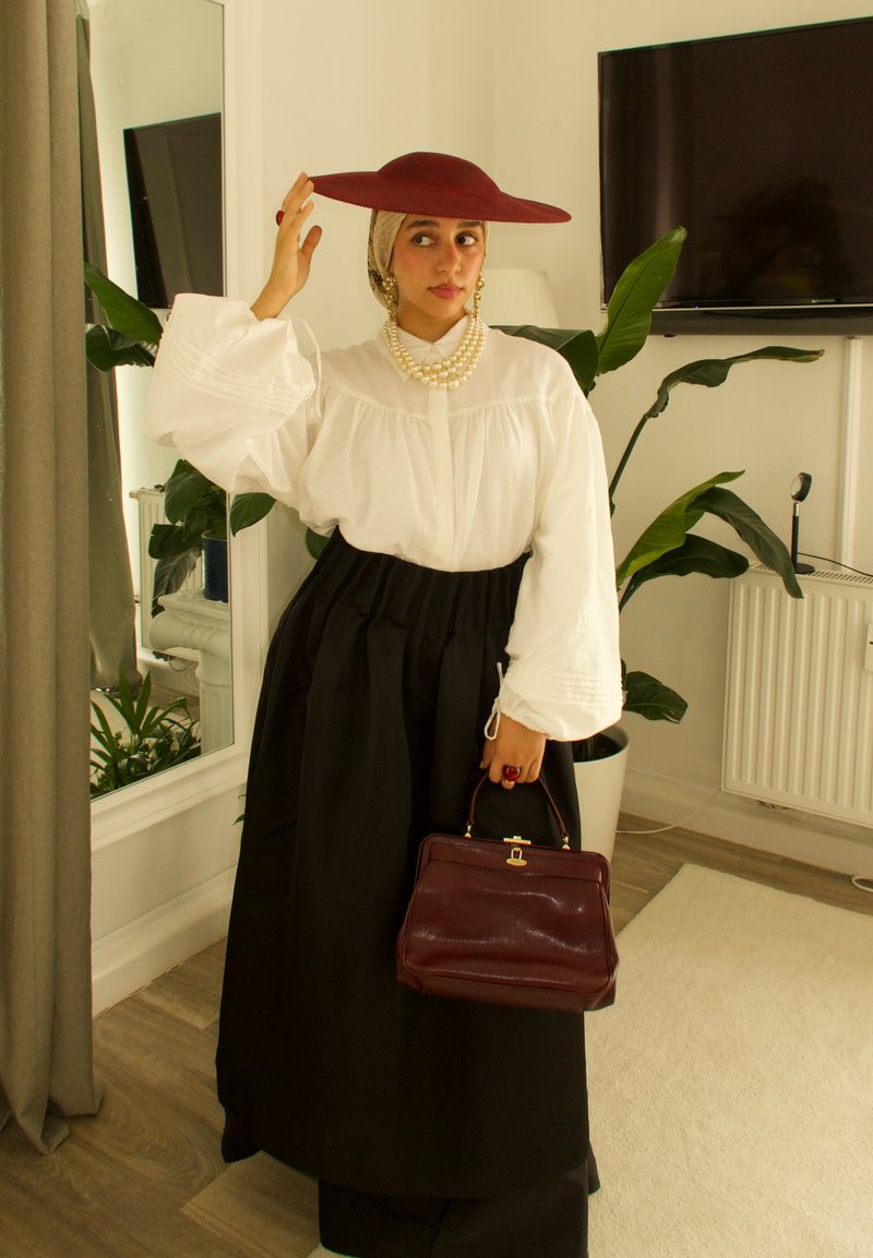 Cappello rosso a falda larga, blusa bianca con maniche a sbuffo, gonna nera e collana di perle. Tiene una borsa bordeaux. Ambientazione interna con piante.