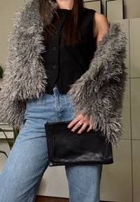 Personne portant un manteau en fausse fourrure grise sur un gilet noir boutonné et un jean bleu, tenant une pochette en cuir noir.