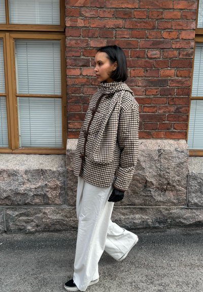 Mujer caminando junto a una pared de ladrillos, vestida con una chaqueta marrón de cuadros, bufanda a juego, pantalones blancos, guantes negros y zapatos negros.