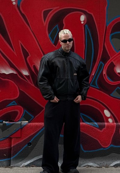 Chaqueta de cuero negra con cuello alto, pantalones anchos negros y gafas de sol frente a un vibrante fondo de graffiti rojo y azul.