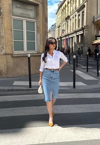 Witte blouse, lichte denim kokerrok met een split aan de voorkant, gele high heels en een kleine witte handtas. Achtergrond van een stadsstraat.