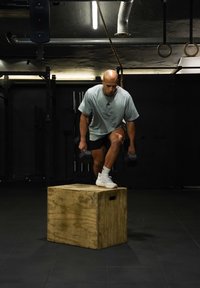 Homme montant sur une boîte en bois en tenant des haltères dans une salle de sport faiblement éclairée avec des anneaux d'exercice et des cordes suspendus au plafond.