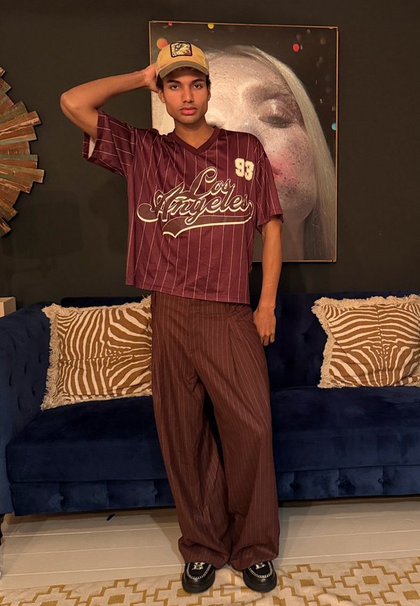 Giovane uomo con un cappellino beige, camicia a righe bordeaux con la scritta "Los Angeles" e pantaloni coordinati in piedi davanti a un divano blu con cuscini stampati a zebra.