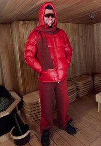 Crvena izolirana puhasta jakna s kapuljačom, odgovarajuće crvene hlače i smeđi šal. Crne cipele i velika crvena sunčana naočala. Drveni unutarnji prostor saune.