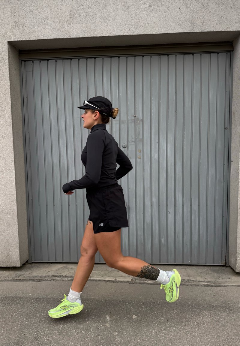 Femme en tenue de sport noire et casquette courant devant une porte de garage grise fermée, portant des chaussures de course jaune fluo et des chaussettes blanches.