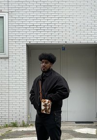 Juodas bomber stilius švarkas, juoda marškinėliai ir kelnės; laiko mažą rudą ir baltą raštuotą per petį nešamą rankinę; stovi šalia baltų plytų sienos.