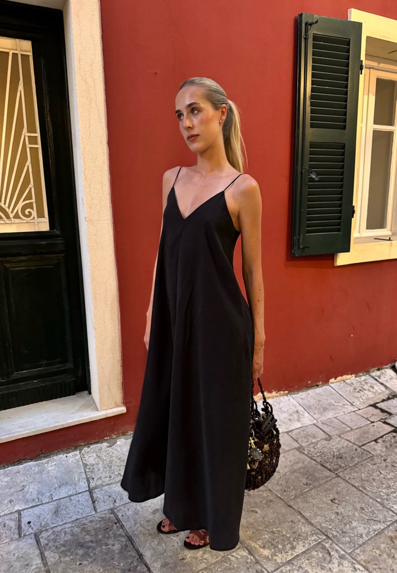 Γυναίκα με ξανθά μαλλιά σε μαύρο αμάνικο φόρεμα στέκεται σε πεζοδρόμιο από πέτρα κοντά σε κόκκινο τοίχο με πράσινα κουφώματα.