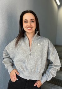 Šedá mikina na zips s vysokým golierom a rebrovaným lemom, na hrudi s malým bielym logom. Čierne nohavice dopĺňajú tento outfit.