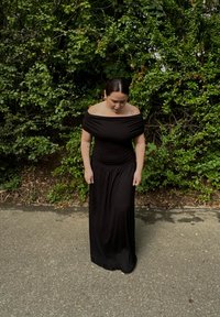 Robe maxi noire à épaules dénudées avec un corsage ajusté et une jupe fluide. Texture lisse, détail froncé à la taille, portée devant un feuillage vert.