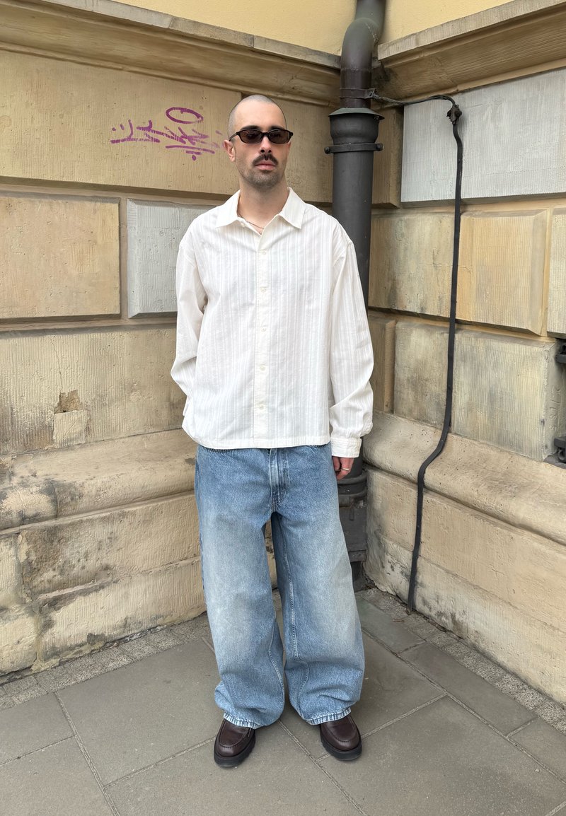 Homme en chemise blanche oversize à boutons, jean ample délavé, chaussures marron et lunettes de soleil, debout contre un mur en pierre beige avec des graffitis.