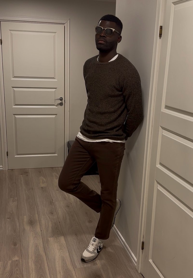 Homme portant des lunettes de soleil, un pull brun foncé, un pantalon marron et des baskets blanches appuyé contre un mur beige entre deux portes blanches fermées.