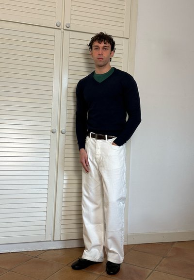 Joven de pie en el interior con un suéter negro, camiseta interior verde, pantalones blancos de pierna ancha, zapatos negros y un cinturón marrón.