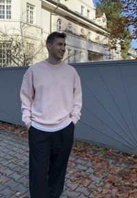 Lichtroze sweatshirt met geribbelde boorden en zoom, gedragen over een wit overhemd, gecombineerd met zwarte broek. Staand tegen een grijs geometrisch hek.