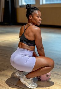 Idrettsbekledning med en svart sports-bh og lavendel høytlivsshortser. En kettlebell ligger på gulvet. Lys, trefarget parkett og naturlig lys.