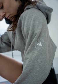 Sweat à capuche gris avec un tissu texturé, présentant un logo blanc sur la manche. Comprend une capuche avec cordon de serrage et une coupe décontractée.