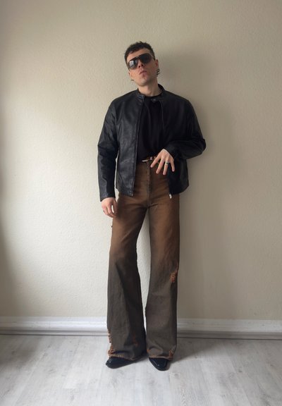 Hombre con gafas de sol grandes, chaqueta de cuero negra, camisa negra, pantalones anchos marrones con detalles desgastados y zapatos negros puntiagudos, de pie en el interior.