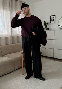 Homme portant un pull à côtes violet foncé et un pantalon noir à jambes larges, tenant un manteau noir. Un bonnet noir et des lunettes complètent le look.