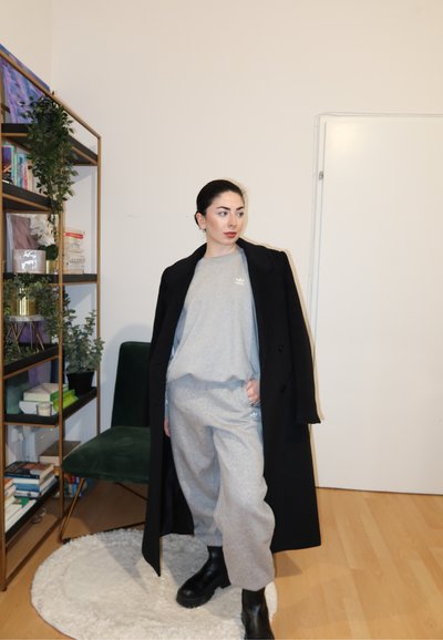 Sudadera gris y pantalones a juego con un logo de Adidas, combinados con un abrigo largo negro. La modelo lleva botas negras y está de pie en una habitación minimalista.