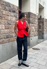 Raudona be rankovių mygtukinė palaidinė su peplum juosmeniu, derinama su tamsiai mėlynomis džinsinėmis kelnėmis, apvyniotomis ties kulkšnimis, ir juodais plokščiais bateliais. Miesto akmeninis fonas.