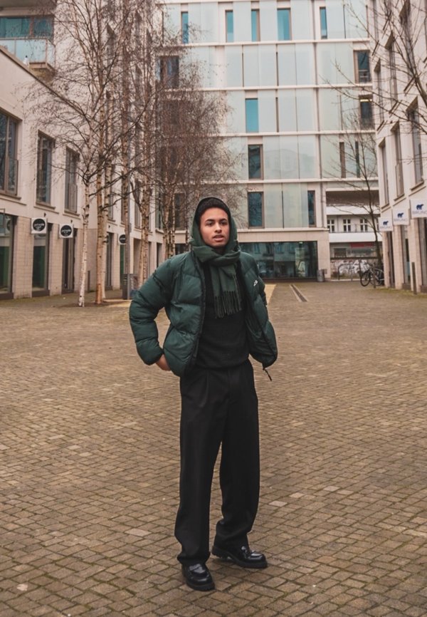 Giacca a vento verde scuro, maglione nero strutturato, pantaloni neri e scarpe nere lucide. Lo sfondo presenta edifici moderni e alberi.