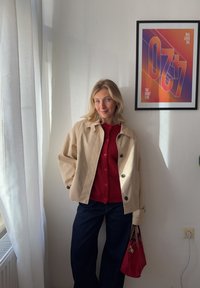 Mulher loira com um casaco bege e um suéter vermelho segura uma bolsa vermelha, em pé junto a uma janela e a um cartaz colorido emoldurado "420 The Right Time".