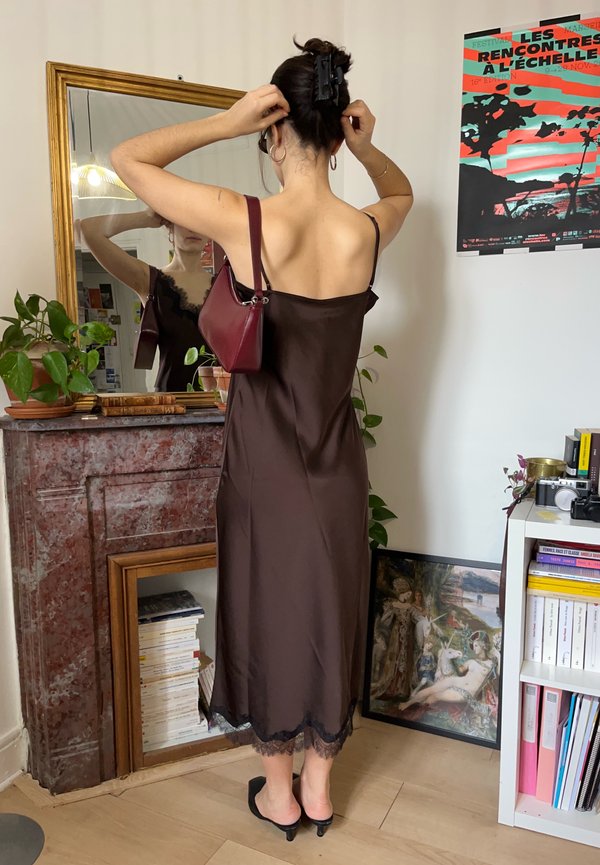 Femeie într-o rochie slip maro își aranjează părul cu o geantă pe umăr, stând în fața unei oglinzi într-o cameră cu plante și cărți.