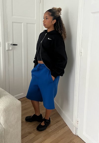 Sudadera negra con cremallera, pantalones cortos azules hasta la rodilla y zapatos negros. Texturas de tela suaves con un diseño deportivo y un ajuste relajado.