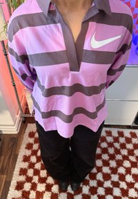 Personne debout, les mains derrière le dos, portant un t-shirt à manches longues rayé rose et marron de la marque Nike et un pantalon noir, sur un tapis à motifs.