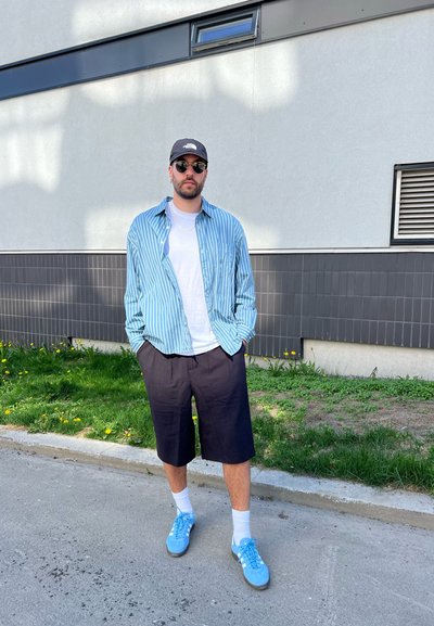 Camisa rayada azul clara y blanca sobre una camiseta blanca, pantalones cortos negros y zapatillas azul claras. Los accesorios incluyen gafas de sol y una gorra.