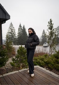 Casaco puffer preto com colarinho alto e textura canelada, combinado com calças escuras e protetores de orelhas, em frente a um fundo natural ao ar livre.