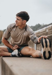T-shirt de sport marron clair avec des accents et un logo gris, associé à un short vert. Baskets noires et grises avec semelles texturées. Posture d'étirement.