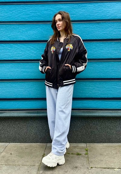 Chaqueta bomber negra con rayas blancas y un diseño de tigre bordado, combinada con pantalones de chándal grises y zapatillas blancas gruesas.
