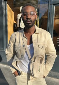 Chaqueta de lona beige con una camiseta sin mangas blanca, pantalones beige y un bolso pequeño con un diseño de la Torre Eiffel verde. Unas gafas claras completan el look.