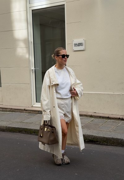 Mujer con un largo abrigo trench beige, camiseta blanca, pantalones cortos con encaje, zapatillas deportivas, gafas de sol y llevando un bolso marrón en una calle de la ciudad.