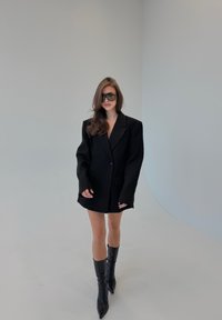 Blazer noir oversize avec épaules structurées, deux poches, fermeture à un bouton, associé à des bottes noires ajustées montant jusqu'aux genoux.