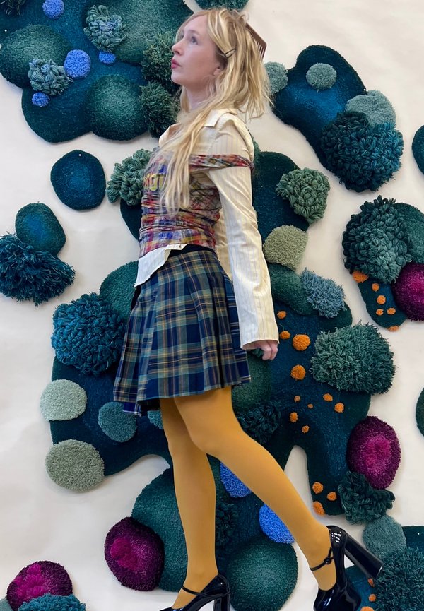 Model draagt een geruite rok in blauw en groen, gele panty's en een aangesloten grafisch topje. De achtergrond toont textuurrijke, kleurrijke stoffen vormen.