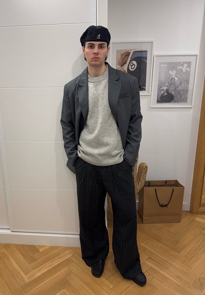 Blazer gris sobre un suéter gris claro, pantalones de pierna ancha a rayas en blanco y negro, y una boina negra. De pie sobre un suelo de madera.