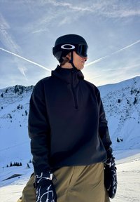Personne portant un casque de ski noir avec lunettes de protection, vêtue d'une veste noire et de gants à motifs, debout sur une pente de montagne enneigée sous un ciel dégagé.