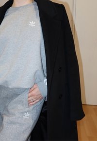 Sweatshirt et pantalon de survêtement gris avec logo Adidas blanc, superposés sous un manteau noir. Texture lisse avec une coupe décontractée et poches latérales.