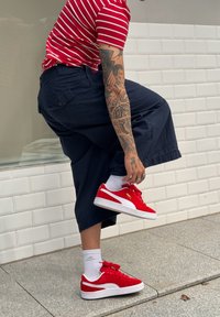 Κόκκινα σουέτ αθλητικά παπούτσια με λευκές λεπτομέρειες, φορεμένα με ναυτικές φαρδιές παντελόνες και μπλούζα με κόκκινες ρίγες. Το φόντο έχει λευκούς πλακίδια τοίχου.