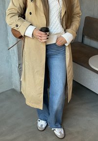 Pessoa vestindo um casaco bege, suéter de malha branca, jeans de perna larga azul e tênis brancos segurando um copo de café dentro de casa.