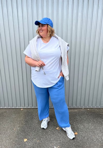 Conjunto casual que incluye una camiseta blanca de manga corta, pantalones sueltos de color azul claro y una gorra azul, con un suéter ligero drapeado sobre los hombros.