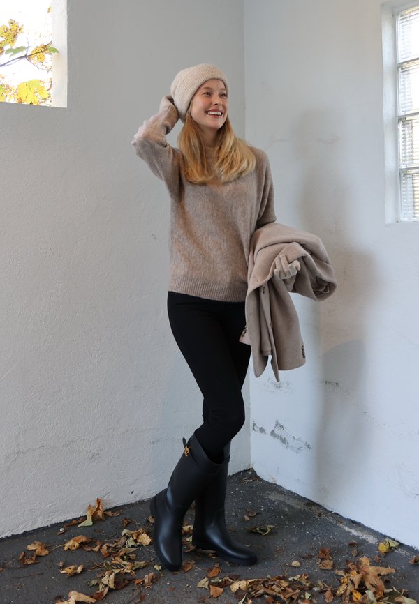 Pull en tricot beige, leggings noirs et bottes en caoutchouc noires. Tenant un manteau marron clair, debout dans un espace intérieur minimaliste avec des feuilles sur le sol.