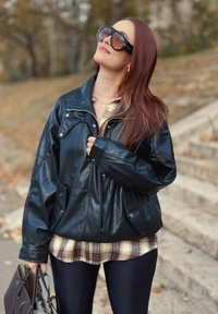 Sort læderjakke med lynlås og trykknaplommer, båret over en ternet skjorte. Modellen holder en brun taske, med mørke leggings og store solbriller.