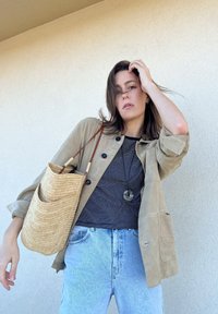 Μπεζ σουέτ μπουφάν με μαύρα κουμπιά, ναυτικό και λευκή ριγέ πουκάμισο, ανοιχτό μπλε τζιν και ένα πλεγμένο ψάθινο τσαντάκι με δερμάτινη λαβή.