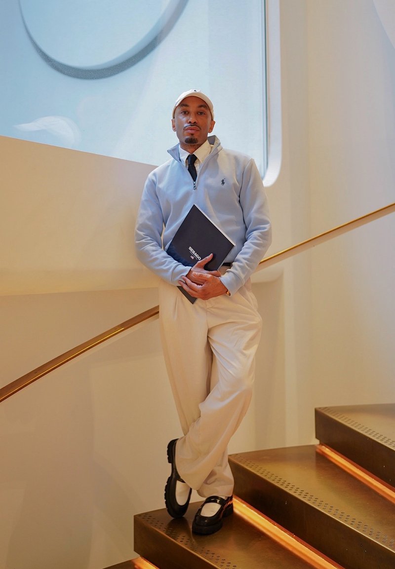 Un homme portant un pull à demi-fermeture éclair bleu clair, un pantalon crème et une casquette blanche s'appuie sur la rampe d'un escalier en tenant un carnet noir à l'intérieur d'un bâtiment moderne.