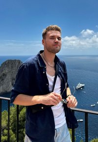 Blue short-sleeve shirt over a white tank top, holding sunglasses. The background features a blue sea and boats, with rocky cliffs and greenery.