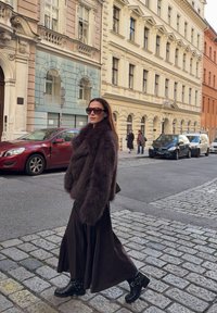 Palton maro din blană artificială, fustă lungă neagră și cizme negre de gleznă cu catarame. Stradă urbană cu piatră cubică și clădiri în stil vintage.