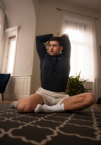 Haut noir à manches longues avec fermeture éclair, shorts blancs et chaussettes blanches. L'individu est assis en tailleur sur un tapis à motifs dans une pièce lumineuse.